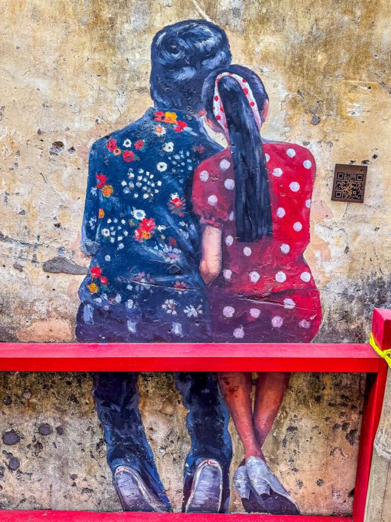 Interaktywny mural w Kwai Chai Hong w Kuala Lumpur, przedstawiający parę siedzącą tyłem na czerwonej ławce na tle starej ściany